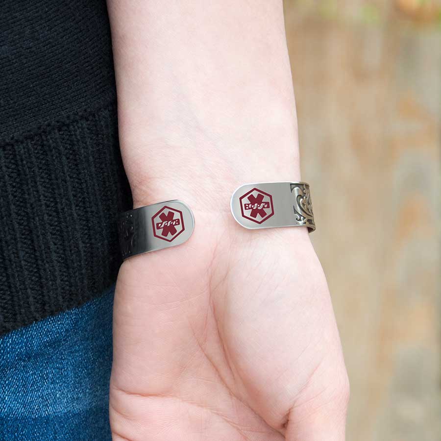 Woman wearing Filigree Medical Alert Cuff. Silver tone stainless cuff. Intricately scrolled design and red caduceus each end