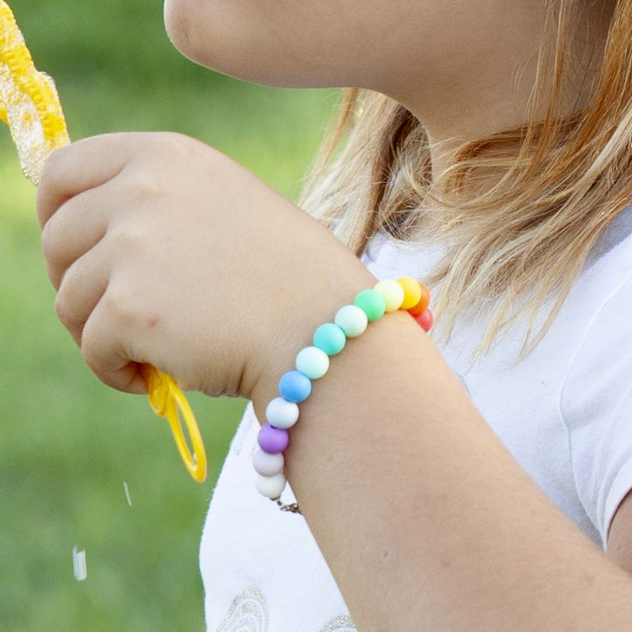 Young girl blowing bubbles in field, wearing medical ID bracelet with rainbow colored silicone beads. 