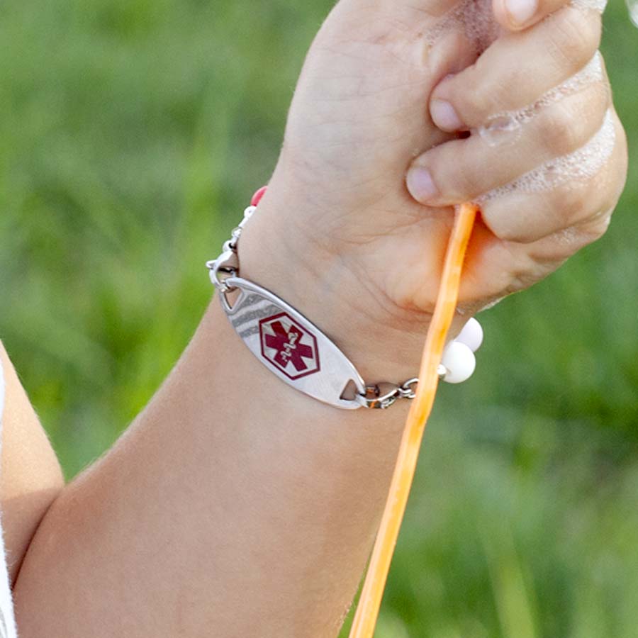 Little girl holding bubble blowing wand, showing medical alert tag on colorful medical ID bracelet with red symbol facing forward.