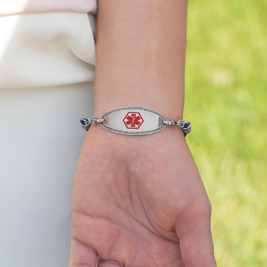 Stainless steel medical ID tag with gray border and red medical symbol worn on navy blue macrame bracelet.