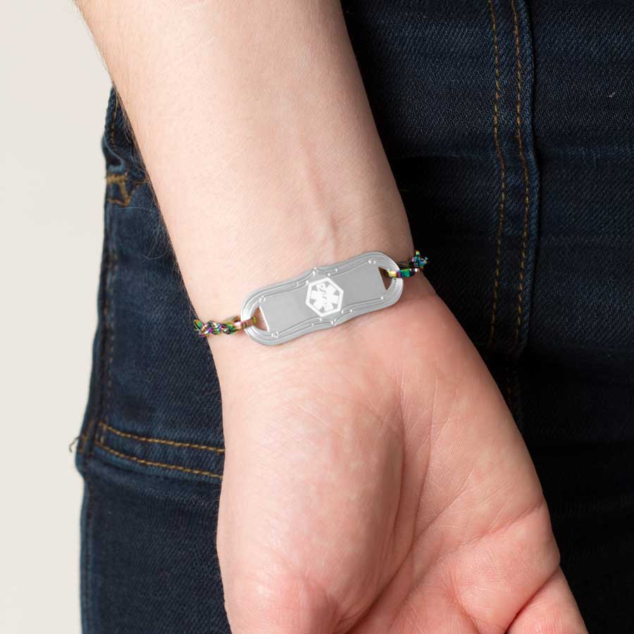 Woman in dark denim wearing multi colored magic finish stainless steel heart link chain bracelet with decorative stainless steel med ID tag with white medical symbol.