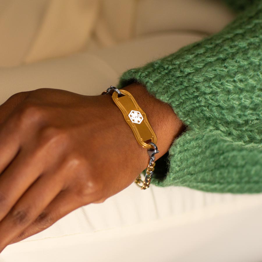 Woman in green sweater and white pants wearing mixed metal figaro chain bracelet with gold tone stainless steel med ID tag with white medical symbol.