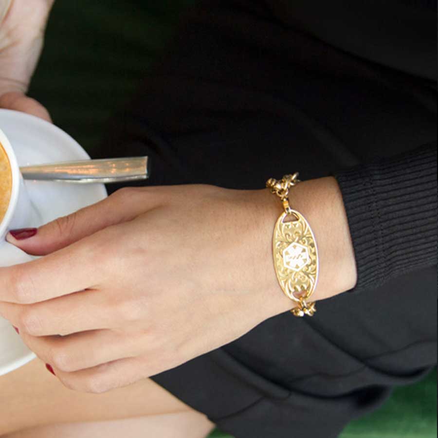 Woman in black top holding coffee wearing three strand gold tone chain bracelet with decorative gold tone stainless steel medical ID tag with white symbol.