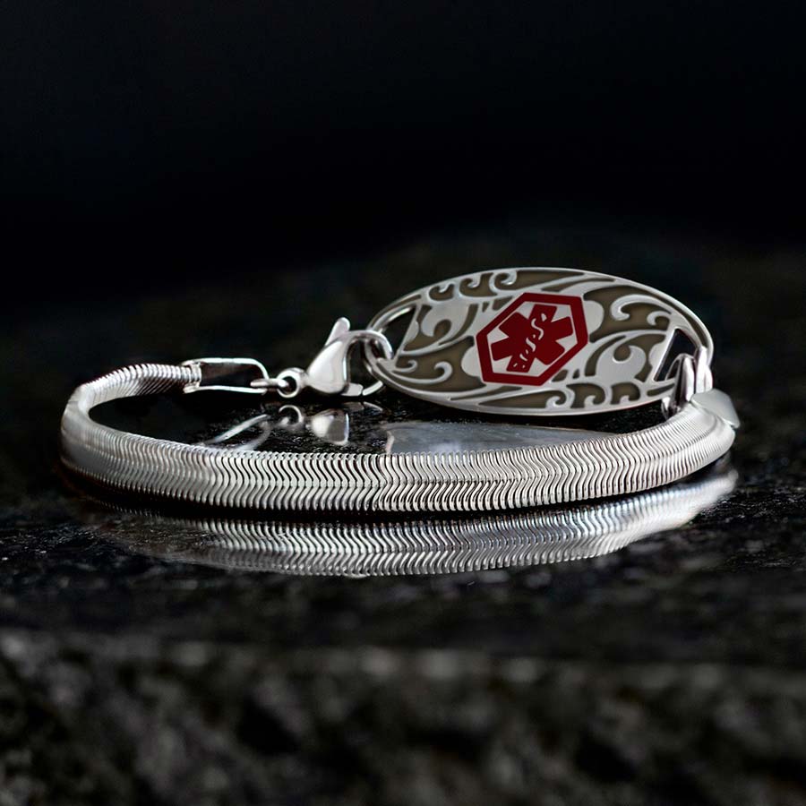 Low angle shot of silver herringbone medical ID bracelet with silver filigree design medical ID tag with red medical caduceus symbol on black stone.