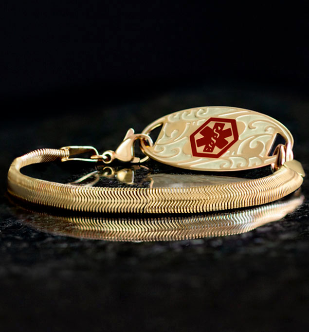 Low angle shot of gold herringbone medical ID bracelet with gold filigree design medical ID tag with red medical caduceus symbol on black stone.