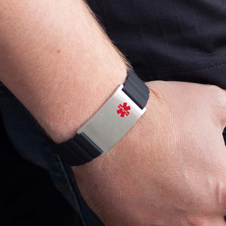 Man wearing blue jeans and black shirt displays magnetic silicone bracelet with black band, silver tag, and red caduceus on his wrist.