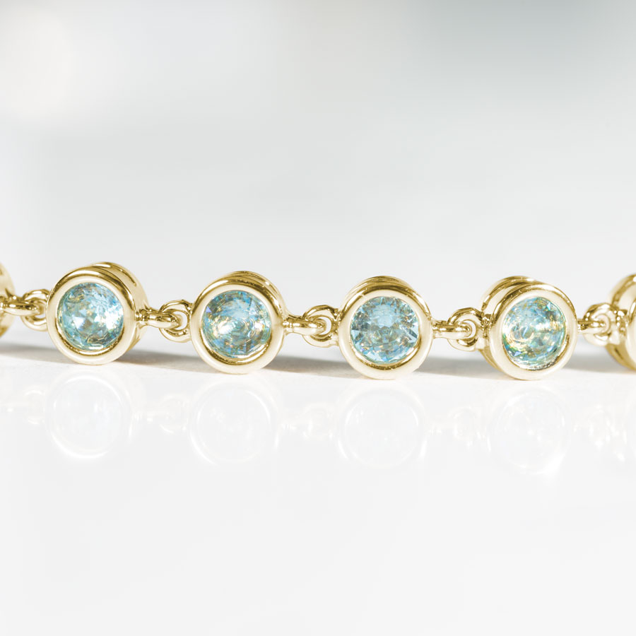 Close up image of gold tennis bracelet with individually linked, round, aquamarine cubic zirconias.