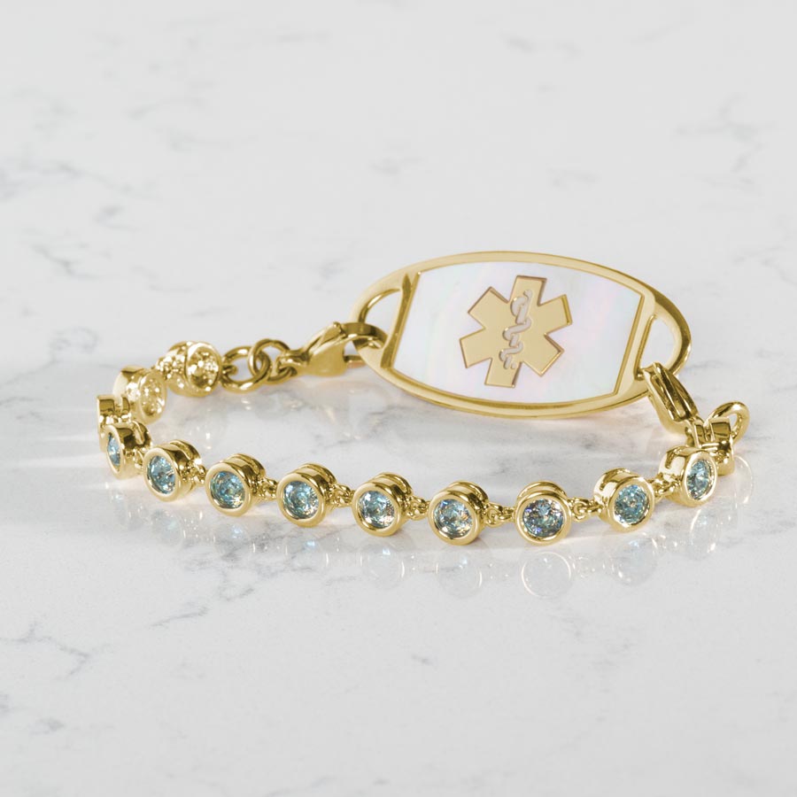 12k gold plated tennis bracelet with individually linked, round, aquamarine cubic zirconias. Attached to a gold tone Mother or Pearl inlaid medical ID tag.