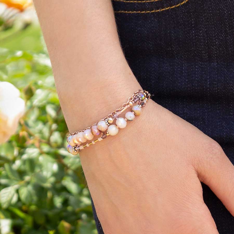 Woman wearing blush and rose gold tone beading bracelet outside with green bush in background.