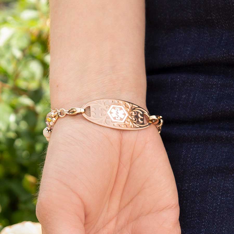 Woman wearing blush and rose gold tone beading bracelet with decorative rose gold medical ID tag outside with green bush in background.