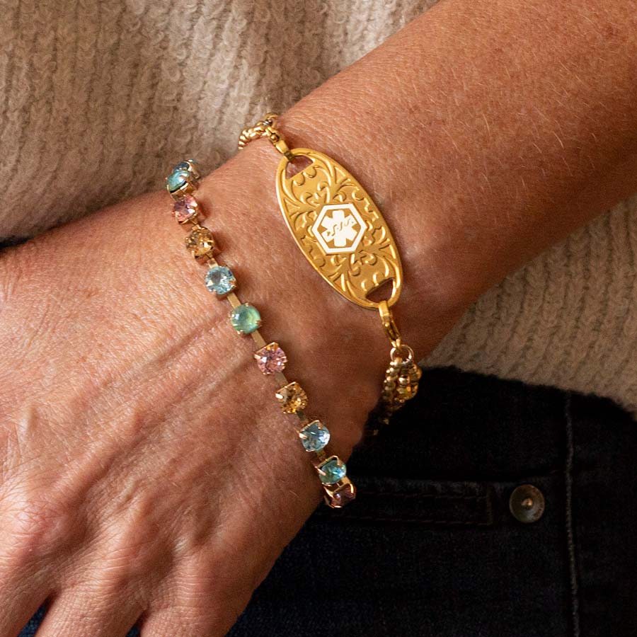 Woman wearing multi colored crystal gold tone box chain bracelet and decorative gold tone stainless steel med ID tag with white symbol.