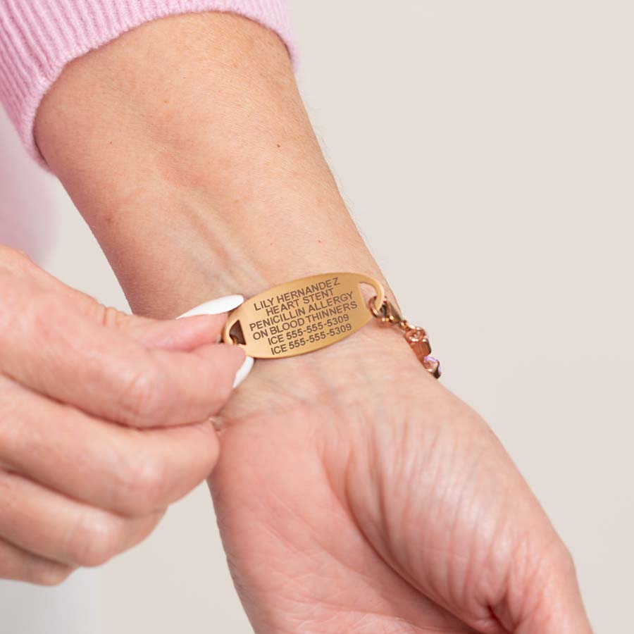 Woman in white pants and pink top wearing rose gold tone chain with lavender crystals and a decorative rose gold tone medical ID tag with premium laser engraving.