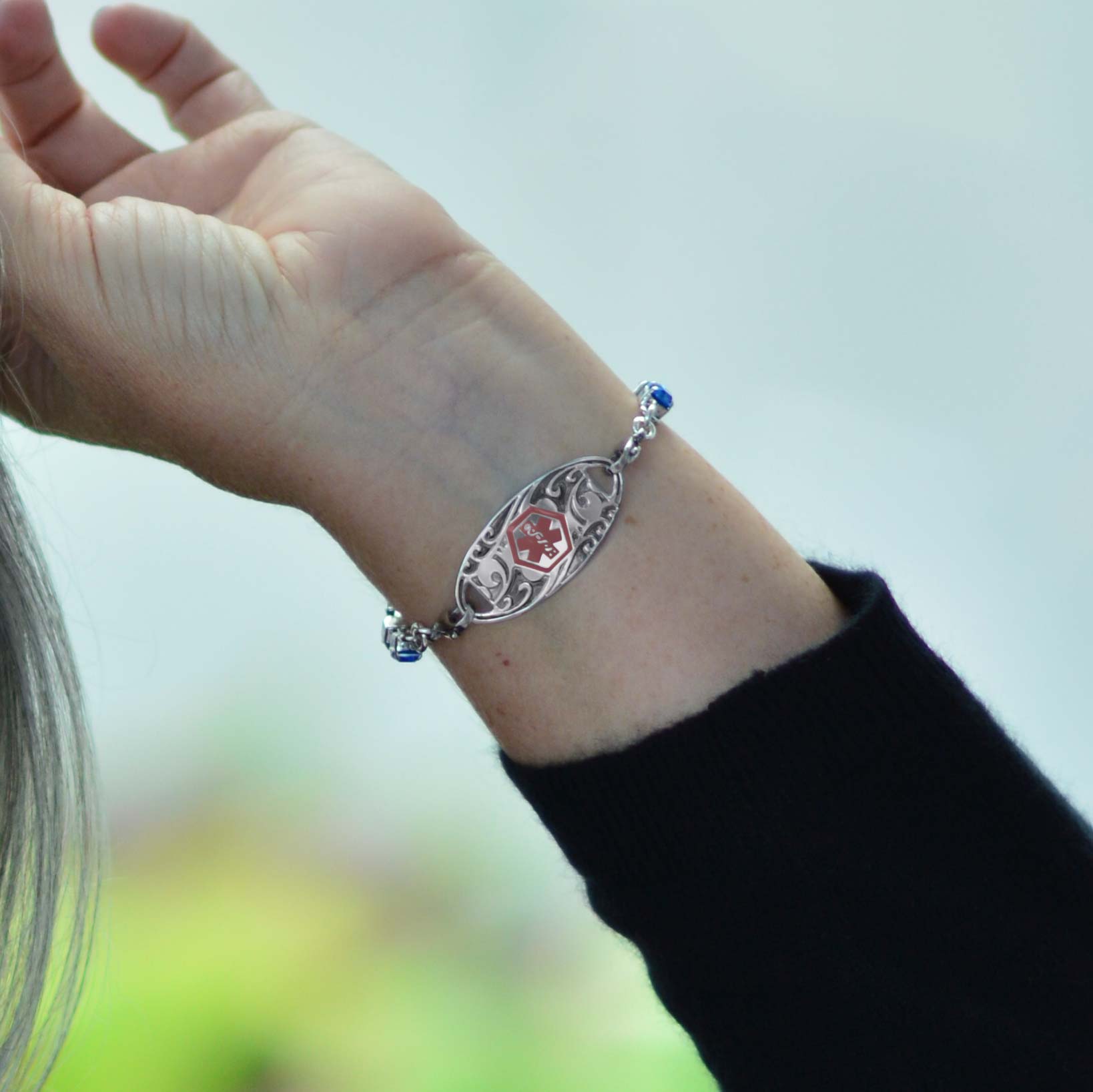 Woman in black top brushing hair from face wearing silver tone box chain bracelet with royal blue crystals and decorative med ID tag.