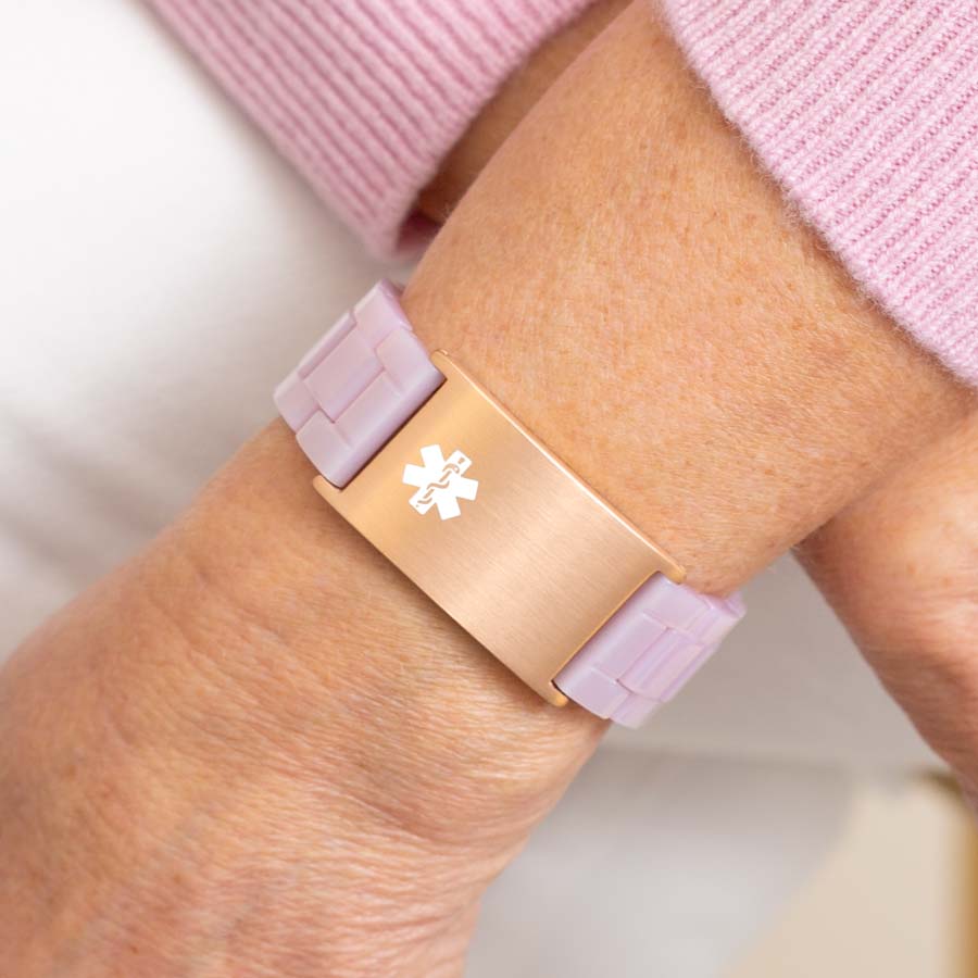 Woman in pink sweater wearing lavender resin linked bracelet with attached rose gold tone stainless steel medical ID tag with off-center white medical symbol.