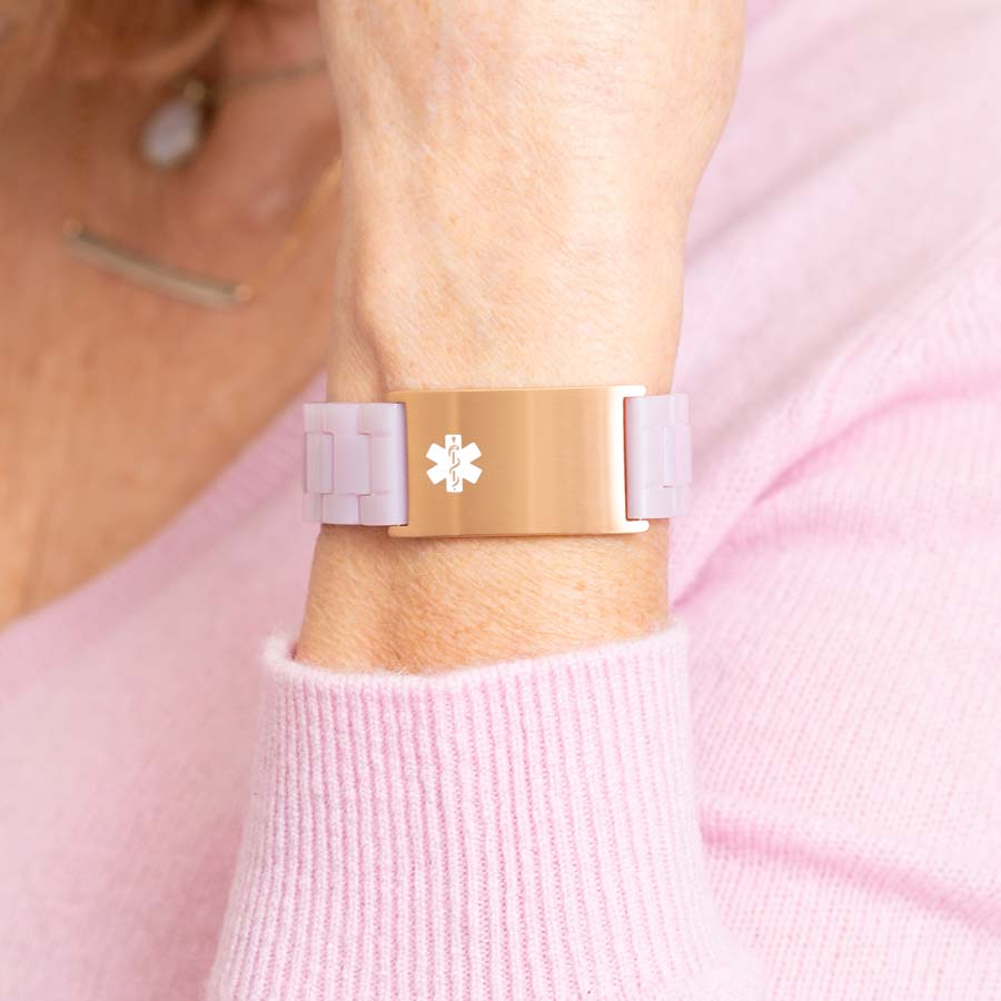 Woman in pink sweater wearing lavender resin linked bracelet with attached rose gold tone stainless steel medical ID tag with off-center white medical symbol.