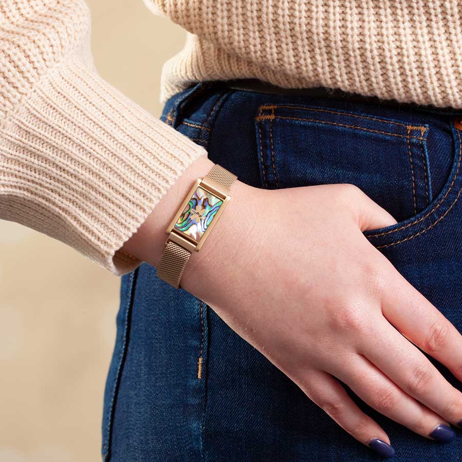 Woman wearing gold tone stainless steel bracelet with adjustable mesh band and affixed ID tag with abalone inlay and gold tone medical symbol.