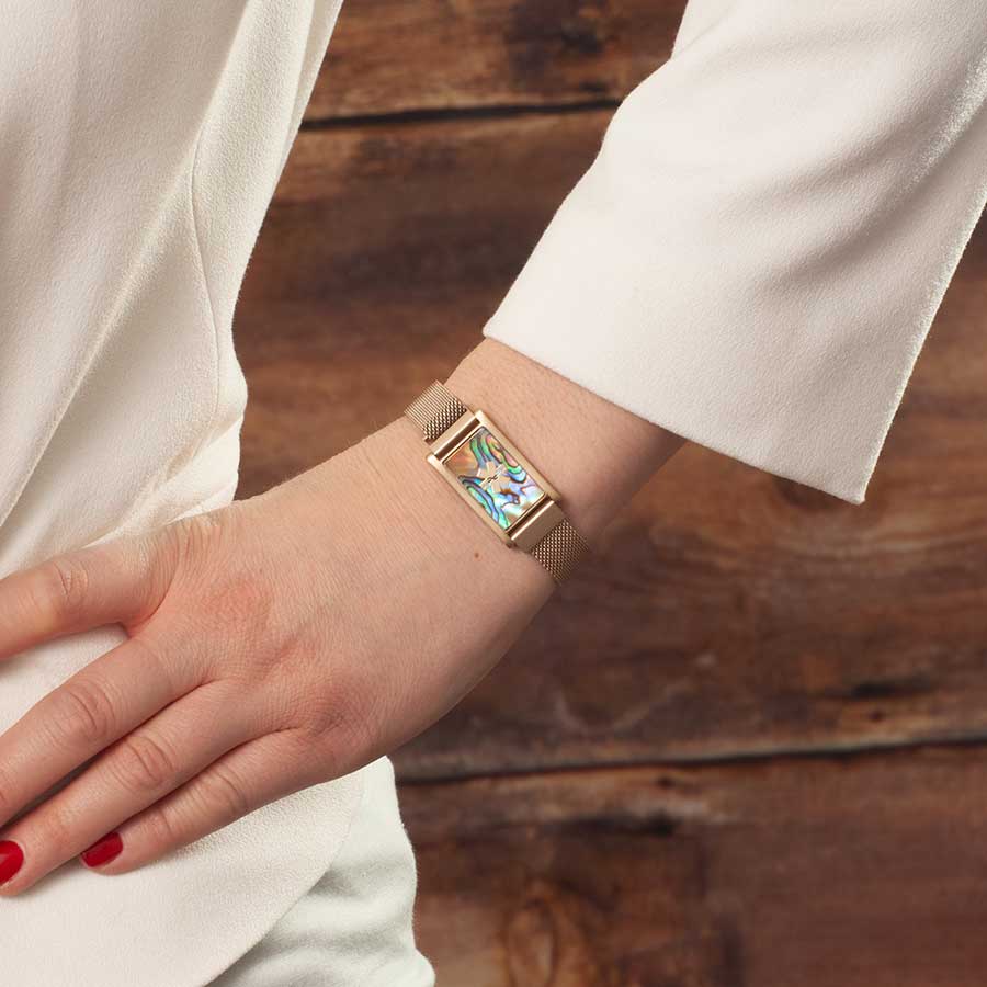 Woman wearing gold tone stainless steel bracelet with adjustable mesh band and affixed ID tag with abalone inlay and gold tone medical symbol.