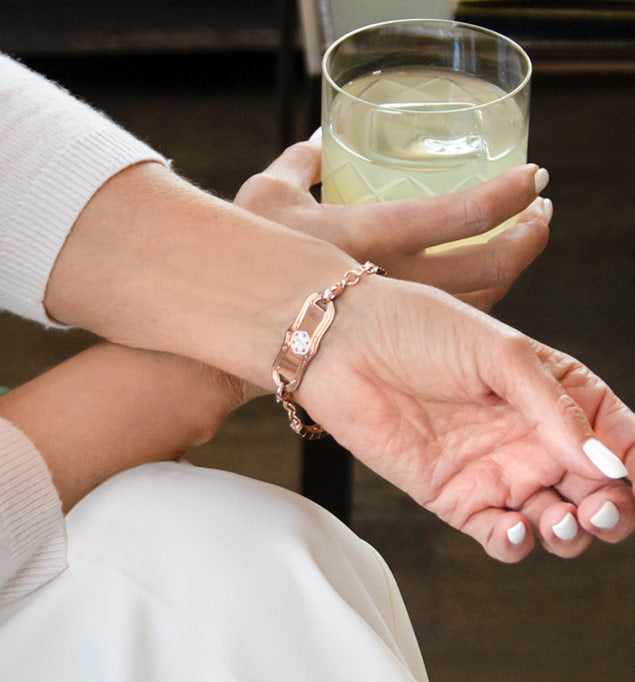 Woman in white holding glass wearing rose gold tone medical ID tennis bracelet showing decorative rose gold tone medical ID tag with white medical symbol.