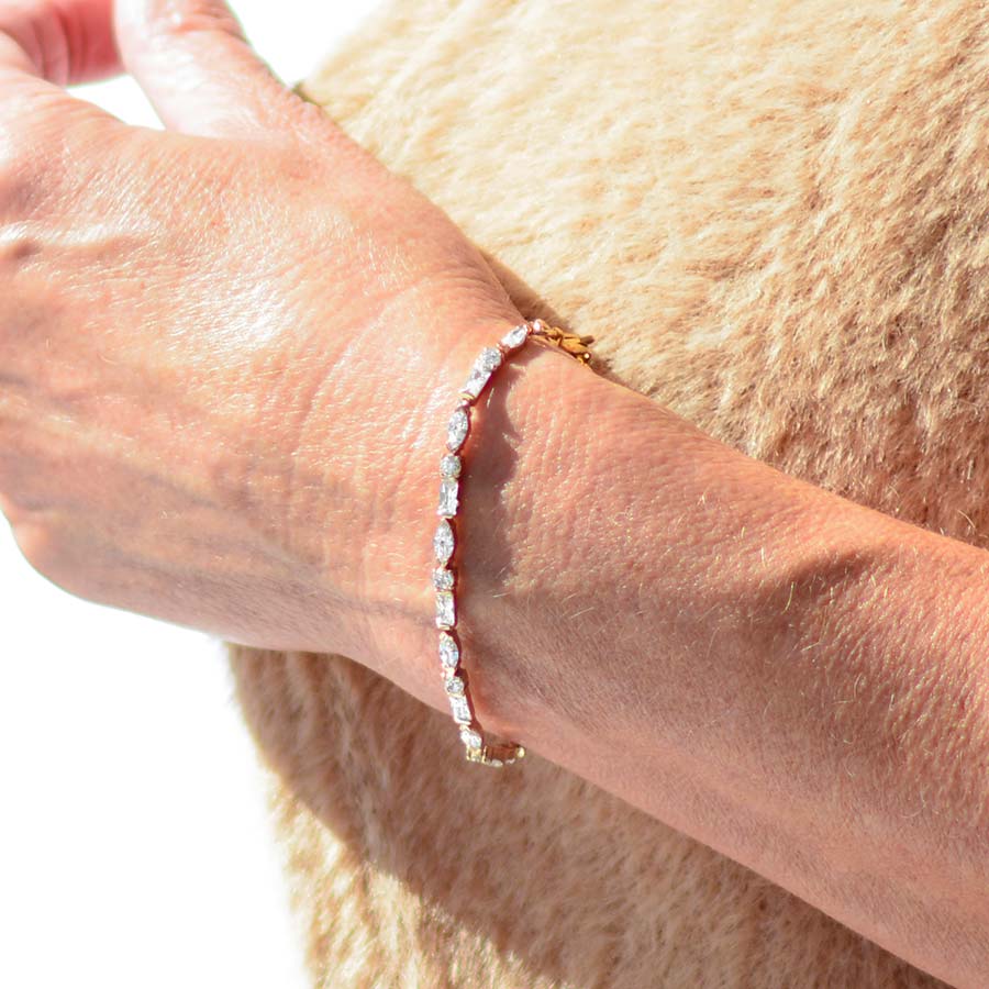 Woman in brown sweater holding collar wearing 12k gold plated tennis bracelet with oval, round, and rectangle cubic zirconia crystals.