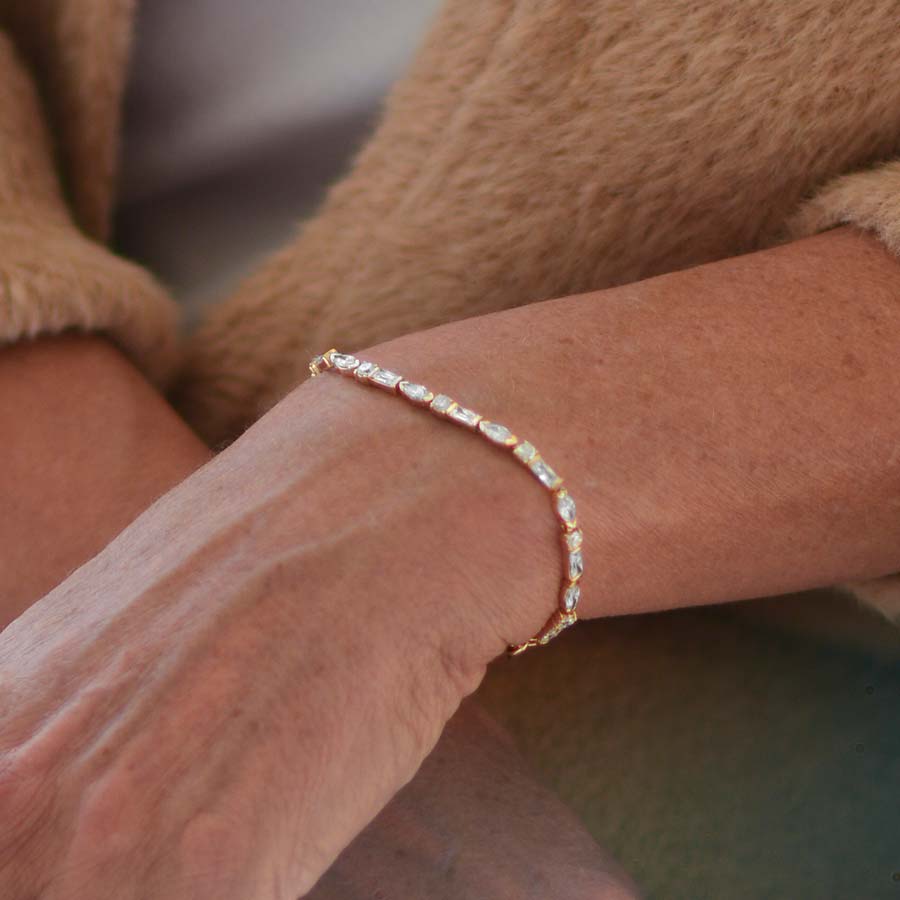 Woman in brown sweater crossing wrists wearing 12k gold plated tennis bracelet with oval, round, and rectangle cubic zirconia crystals.