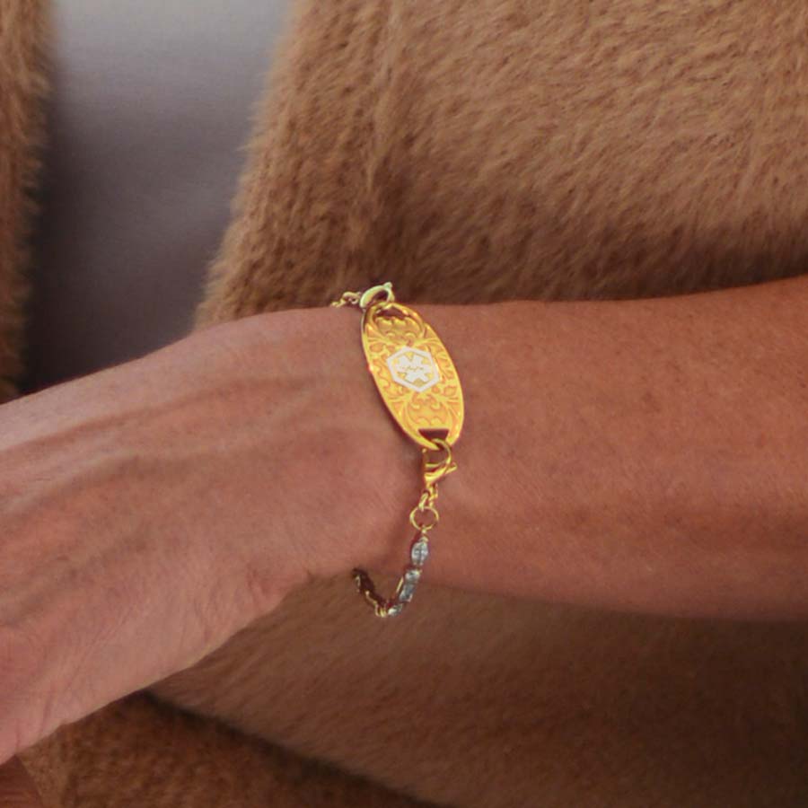 Woman in brown sweater crossing wrists wearing 12k gold plated tennis bracelet with decorative gold tone stainless steel med ID tag.