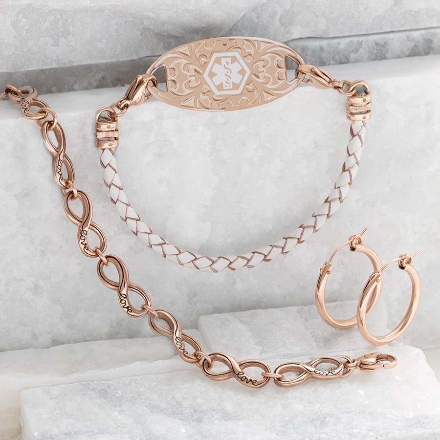 Cream and rose gold braided leather bracelet, rose gold infinity bracelet, rose gold earrings, and decorative med ID tag on granite slabs.