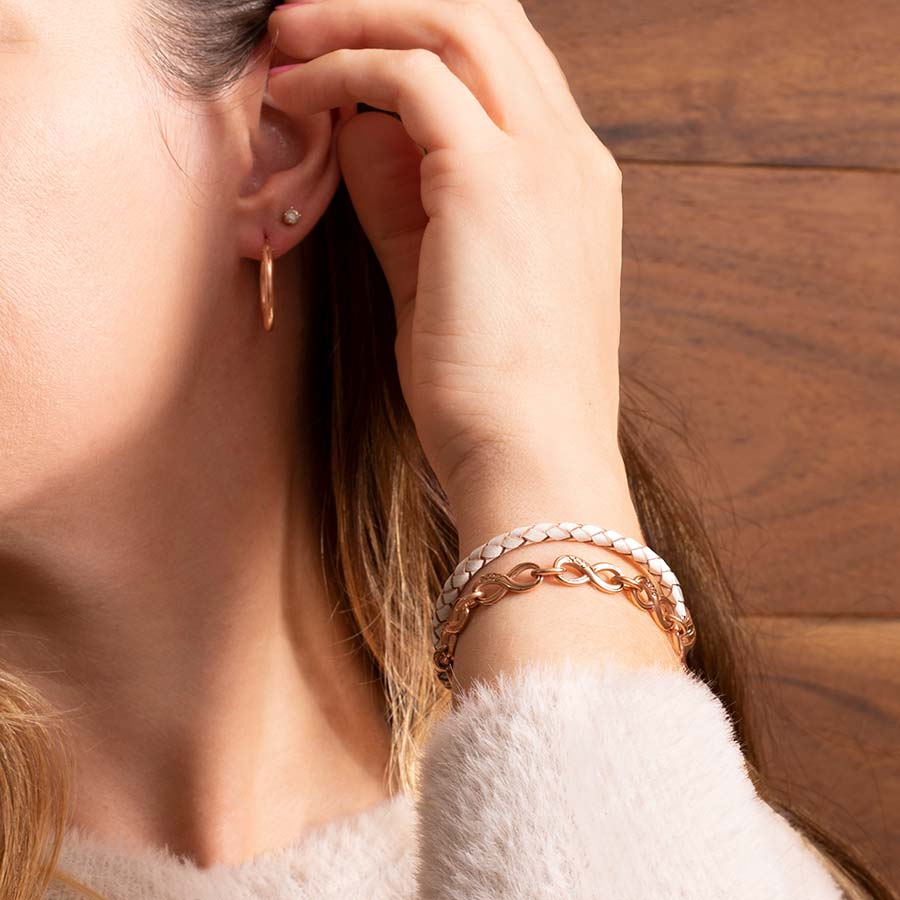 Woman pulling hair behind her ear wearing cream and rose braided leather bracelet, rose gold infinity bracelet, and rose gold hoop earrings.