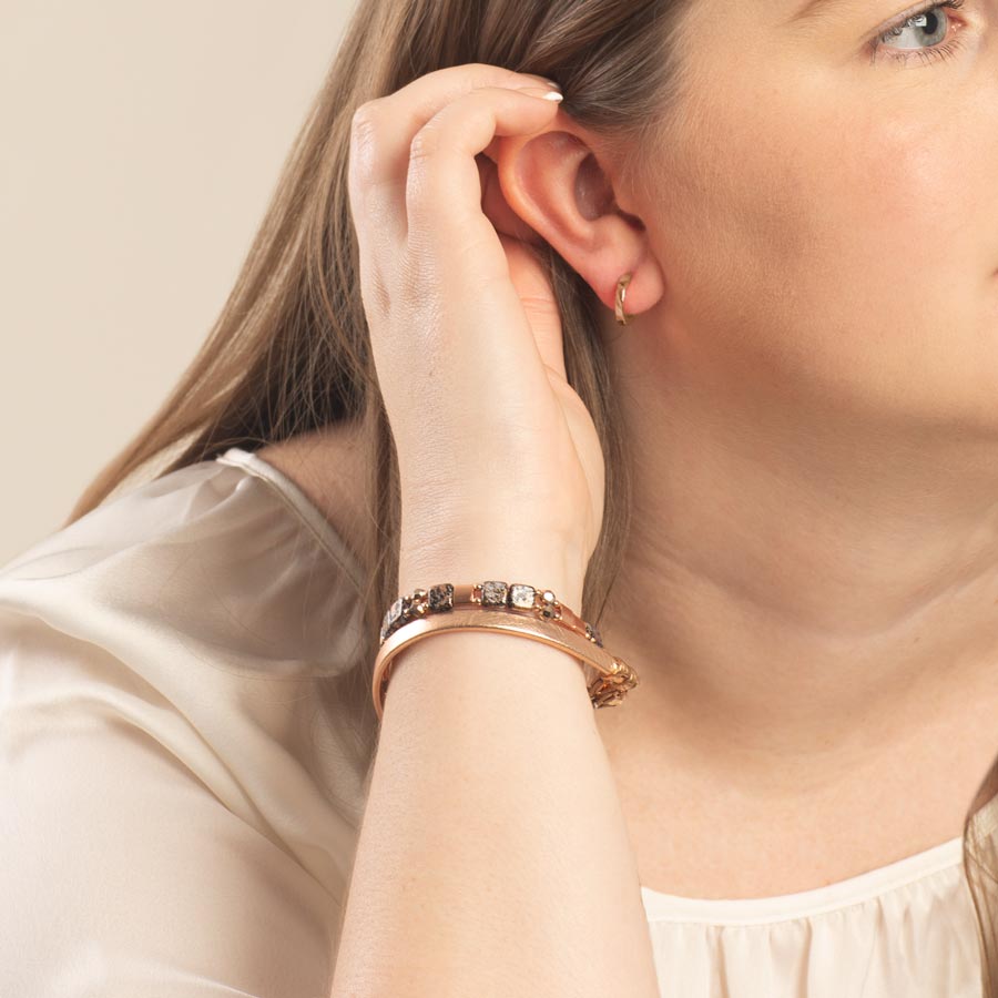 Woman tucking hair behind ear, wearing rose gold hoop earrings; black, rose gold, and crystal beaded stretch bracelet and rose gold paperclip chain bracelet.