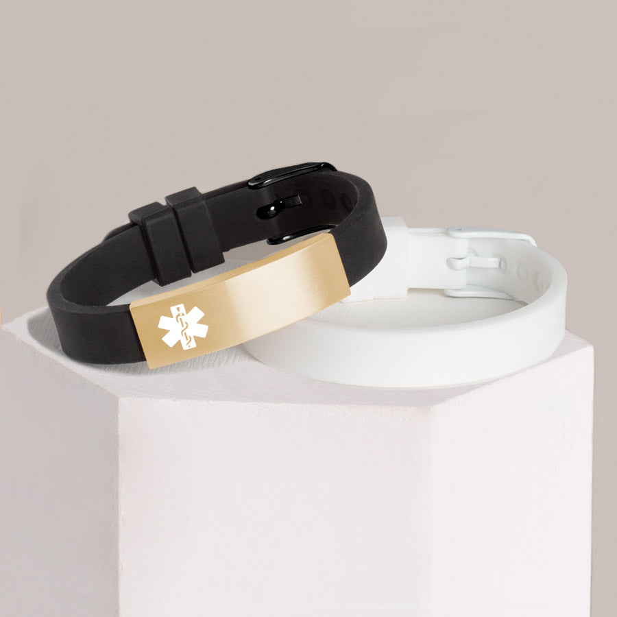 Black silicone medical ID bracelet with gold tone stainless steel medical alert tag stacked with white silicone medical ID bracelet on hexagon block.