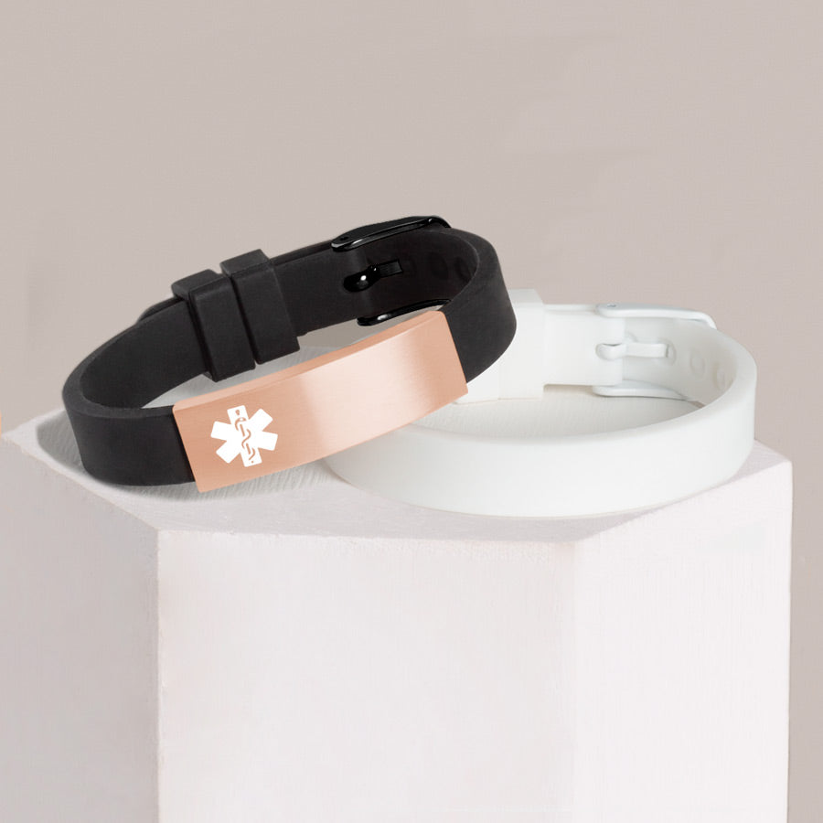 Black silicone medical alert bracelet with rose gold tone stainless steel tag and white silicone medical ID bracelet stacked on white hexagon block.