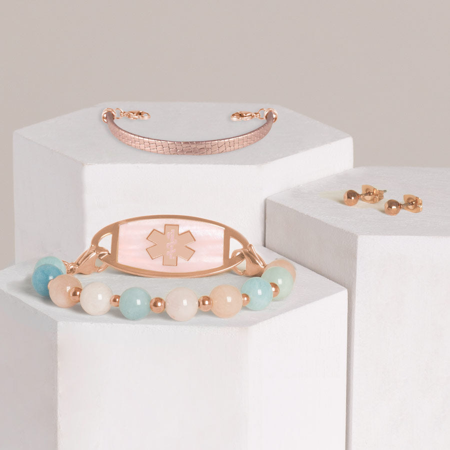 Pink, white, and blue beaded medical ID bracelet paired with rose gold tone stainless steel pink mother of pearl ID tag stacked with rose gold metallic leather medical ID bracelet and faceted, rose gold tone earrings on hexagon blocks.