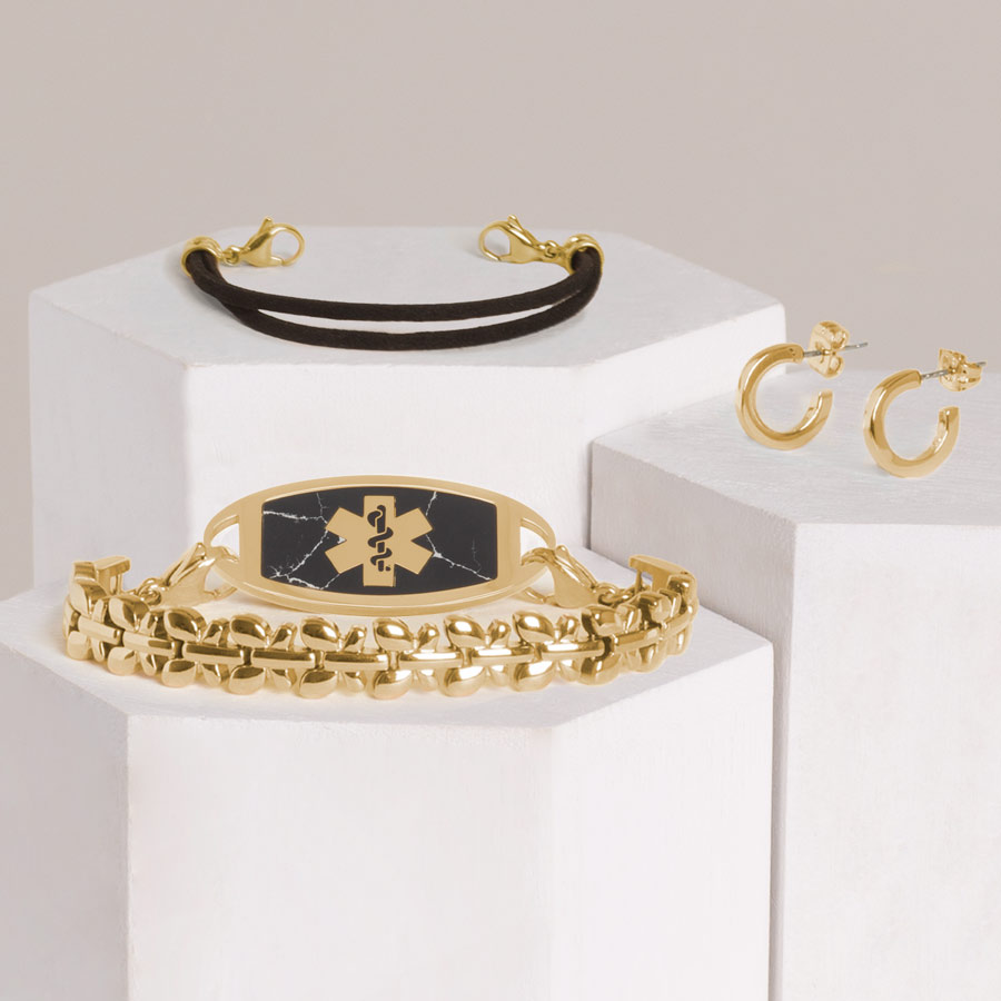 Gold tone stainless steel butterfly shaped bracelet with gold tone stainless steel black marble inlay medical ID tag stacked with black double stranded suede bracelet and gold tone hoop earrings on hexagon block. 