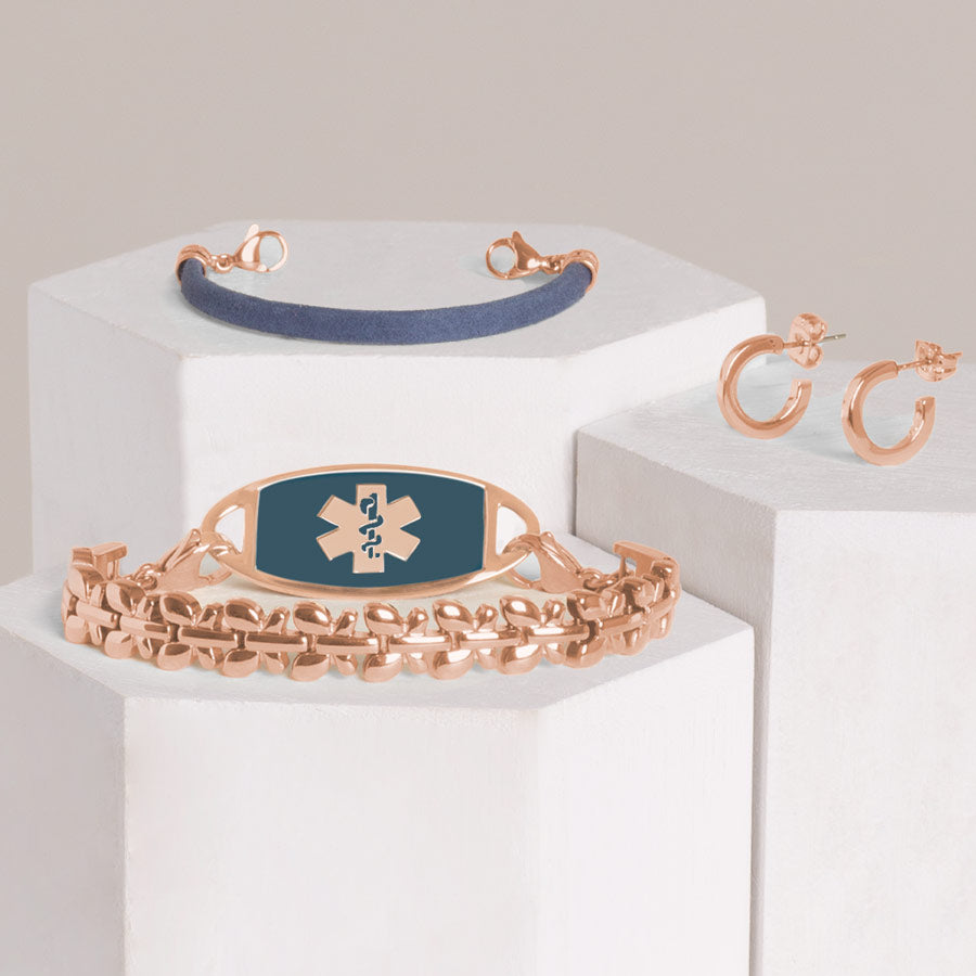 Rose gold tone abstract butterfly medical ID bracelet with rose gold tone sapphire colored inlay ID tag stacked with navy blue leather bracelet and rose gold tone huggie hoop earrings on hexagon blocks.