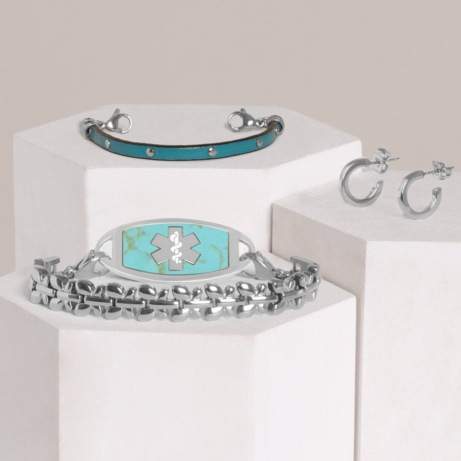 Stainless steel butterfly link bracelet with stainless steel abalone inlay medical ID tag stacked with teal blue leather bracelet with silver studs and silver tone hoop earrings on hexagon blocks.