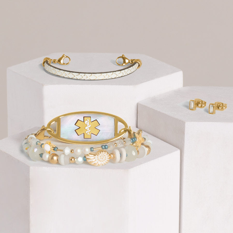 Crystal and pearl beaded medical ID bracelet, cream leather bracelet with scallop textured and gold flakes, and gold tone baguette earrings stacked on hexagon blocks.