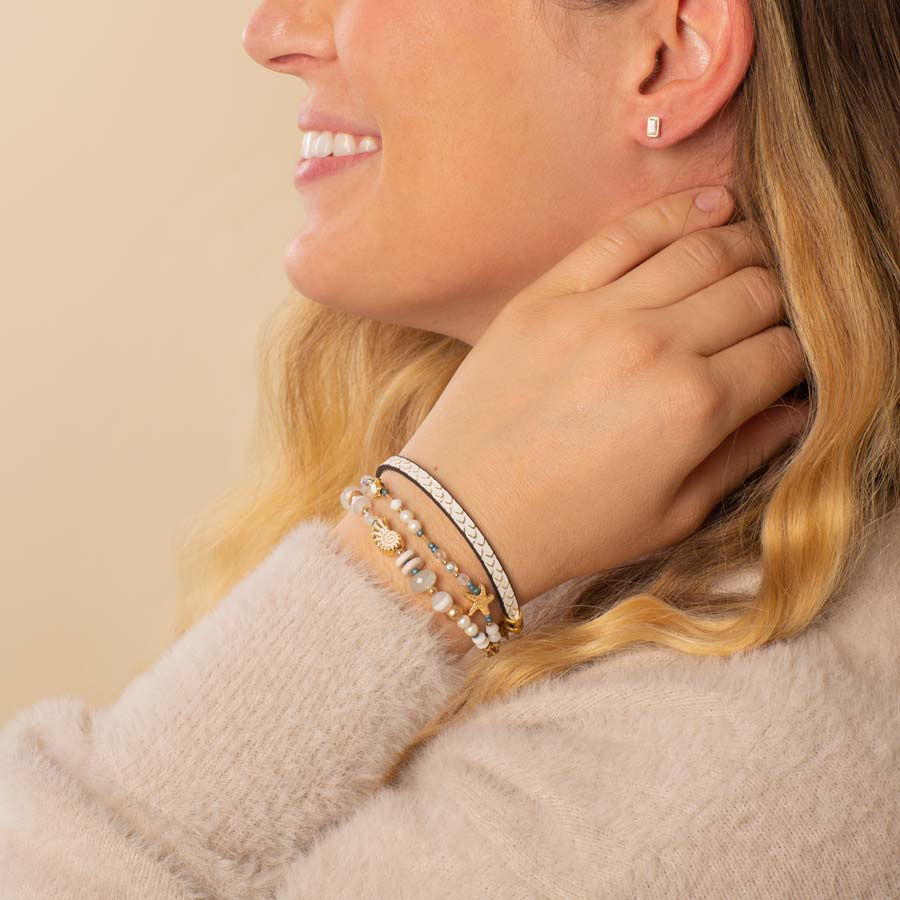 Woman wearing crystal and pearl beaded bracelet layered with cream leather textured bracelet with gold flakes and gold tone baguette earrings.