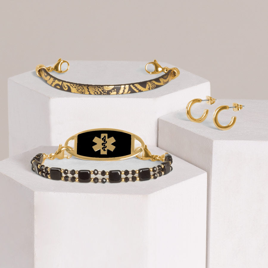 Black and gold beaded stretch bracelet with gold tone stainless steel black inlay ID tag stacked with black leather bracelet with gold paisley pattern and gold tone hoop earrings on hexagon blocks.