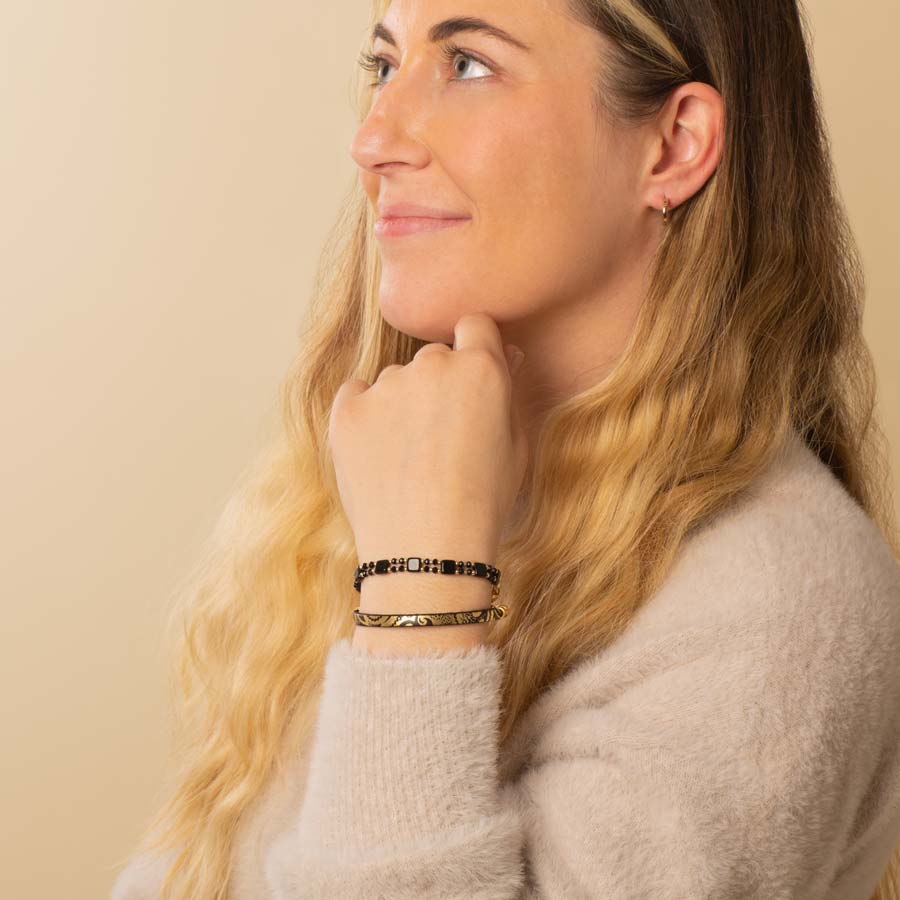 Woman wearing gold tone hoop earrings with black and gold beaded stretch bracelet layered with black leather bracelet with gold paisley pattern.