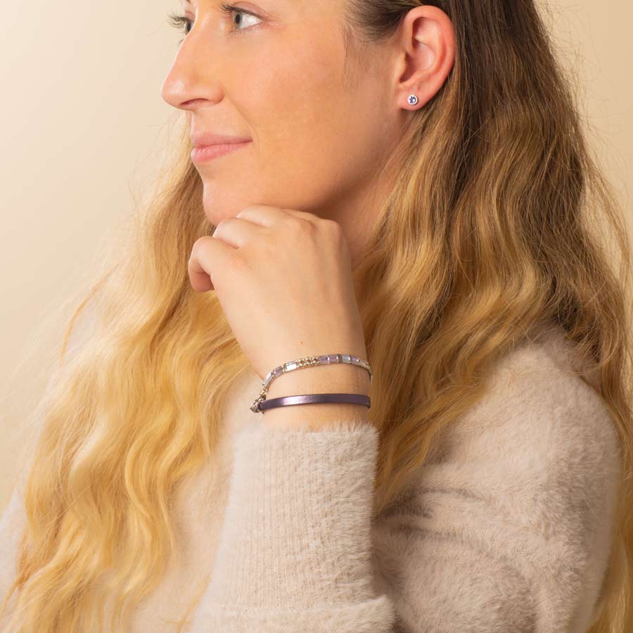 Woman wearing pastel beaded stretch medical ID bracelet layered with purple metallic leather bracelet and silver tone, round tanzanite earrings.