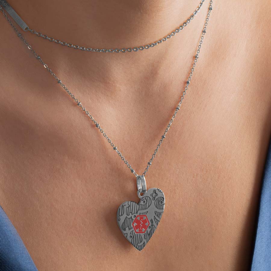 Woman wearing silver heart pendant necklace with decorative pattern and red medical symbol worn on layered necklace strands. 