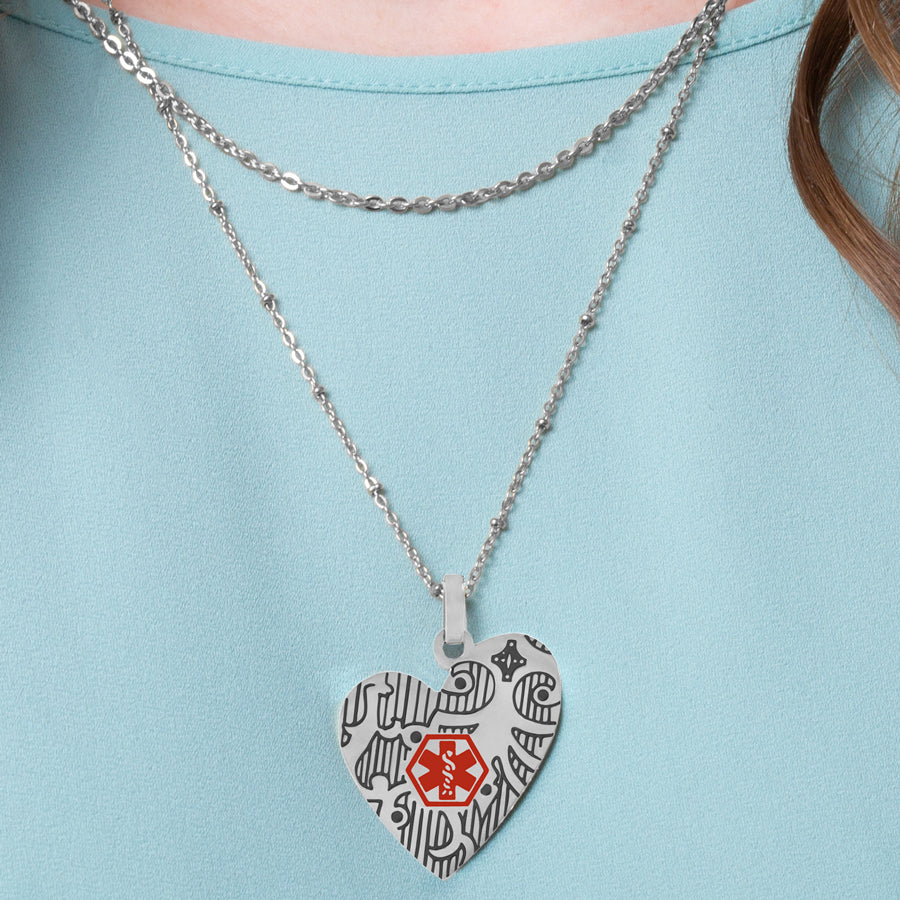 Woman wearing silver heart pendant necklace with decorative pattern and red medical symbol worn on layered necklace strands. 